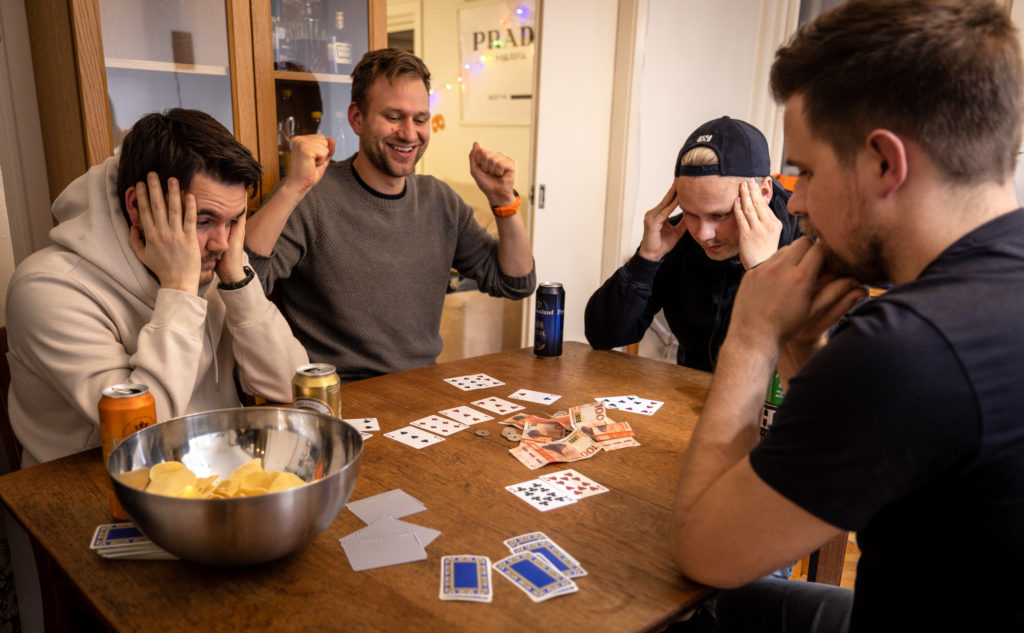 En guttegjeng spiller poker rundt spisebordet. Den ene feirer seieren med henda i været og et stort glis om munn, mens de andre tre guttene sitter med fortvilede utrykk.