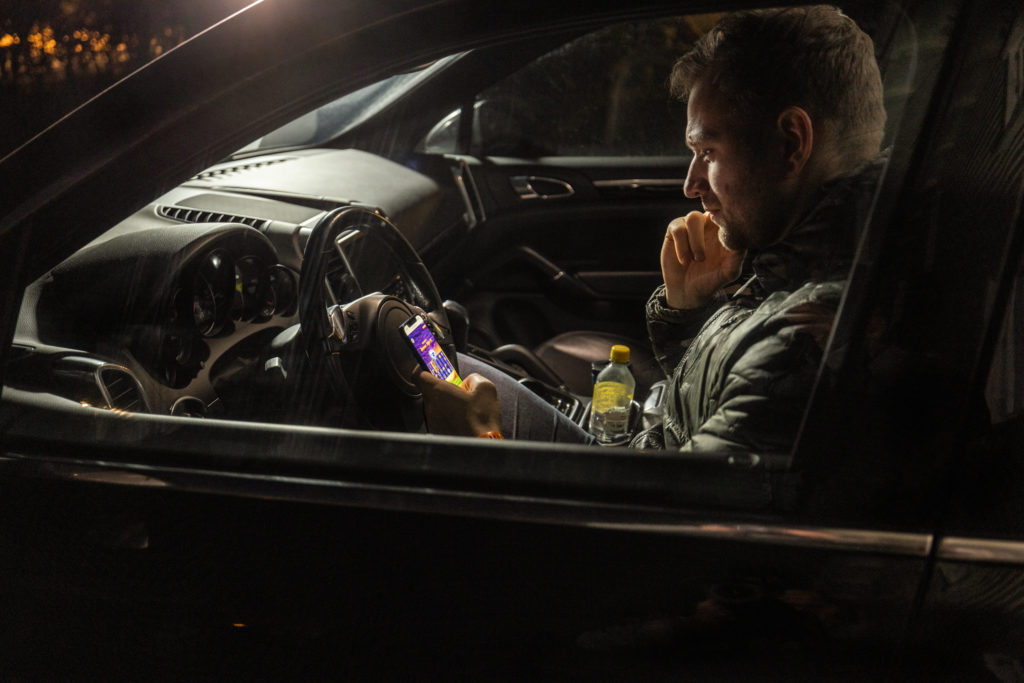En mann sitter i bilen og spiller pengespill på mobilen med et konsentrert utrykk i ansiktet.