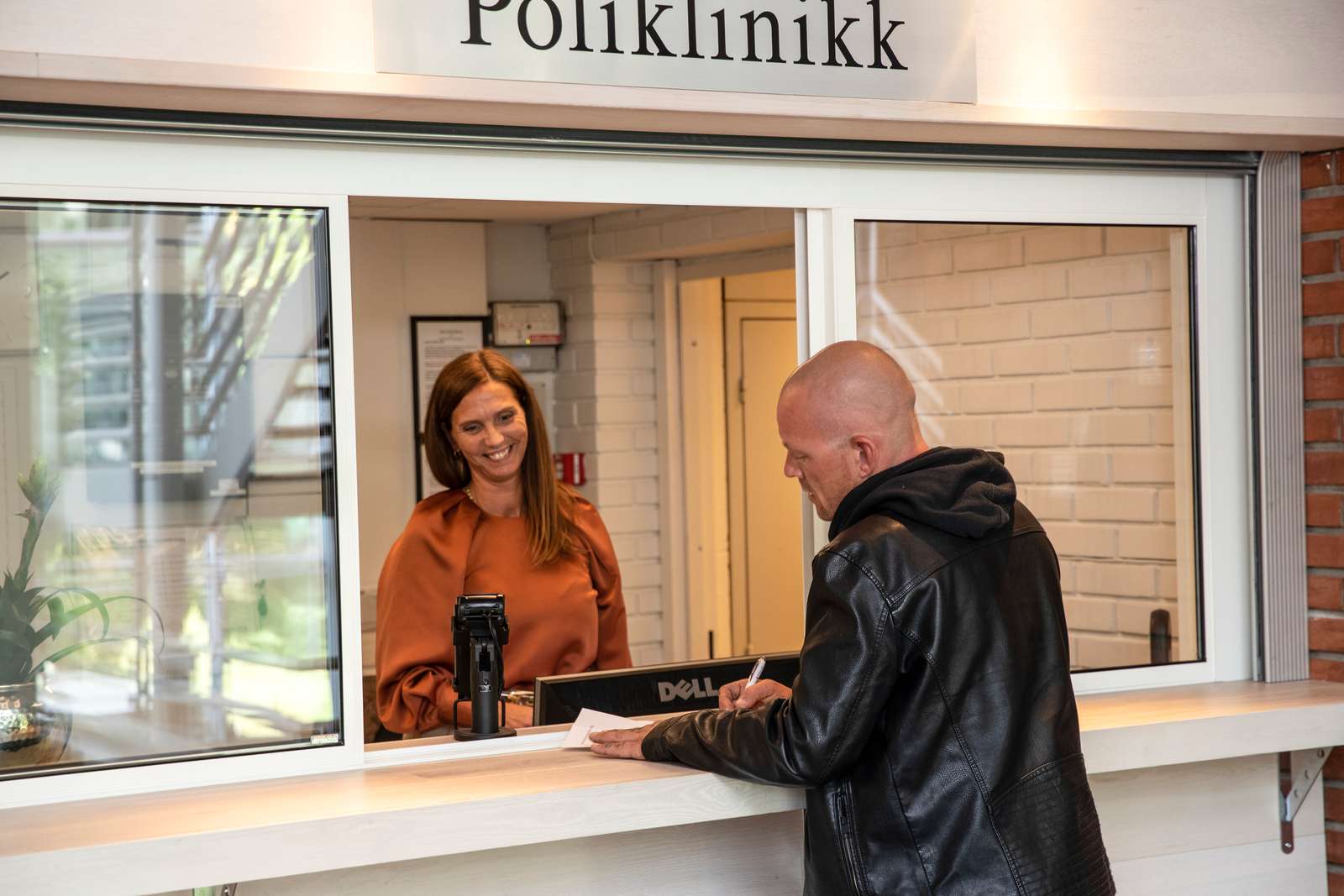 En dame i resepsjonen til Poliklinikken smiler, og tar imot en mann som noterer på et ark