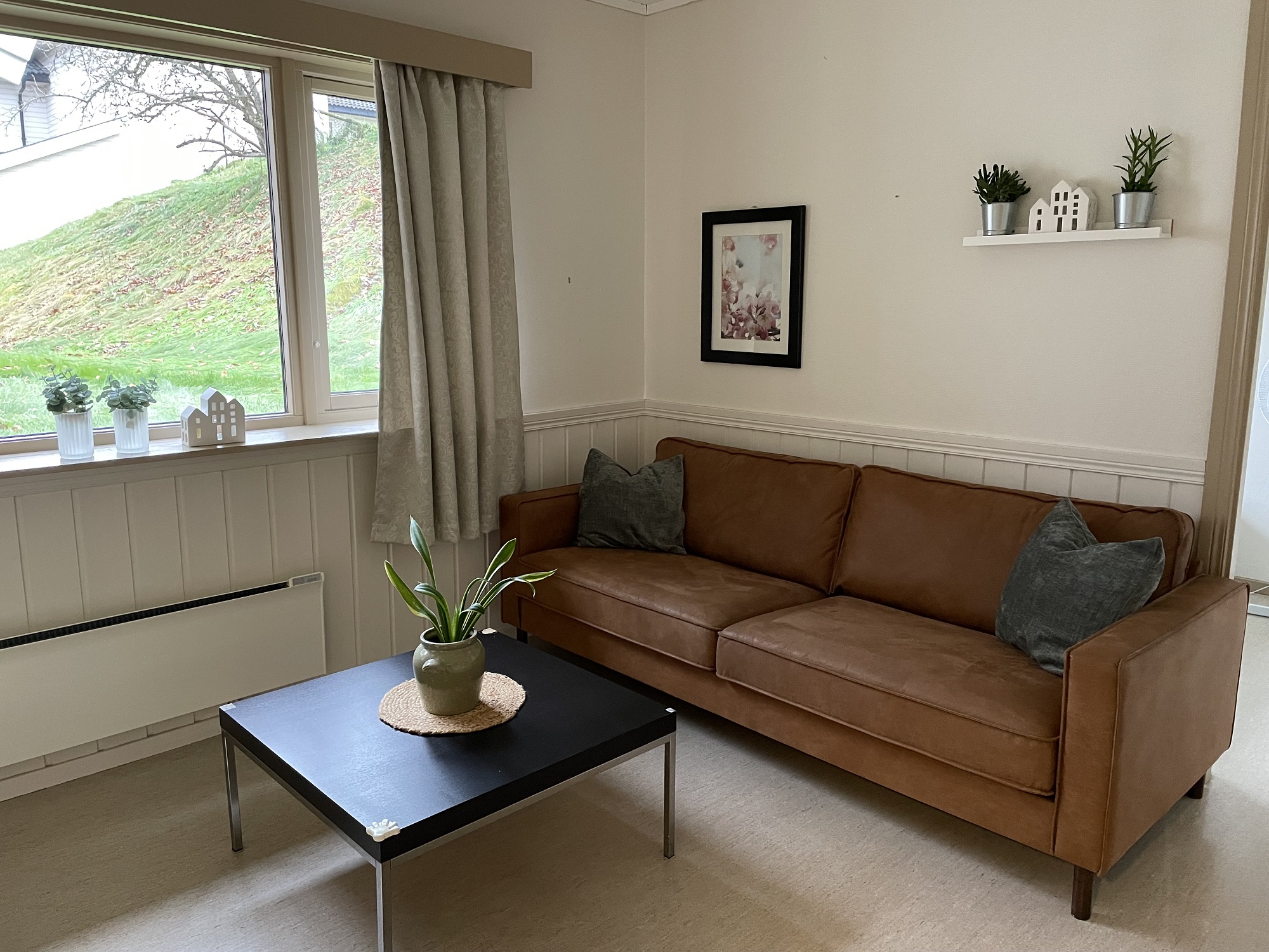 Del av stue med brun sofa, bilder og bord med plante