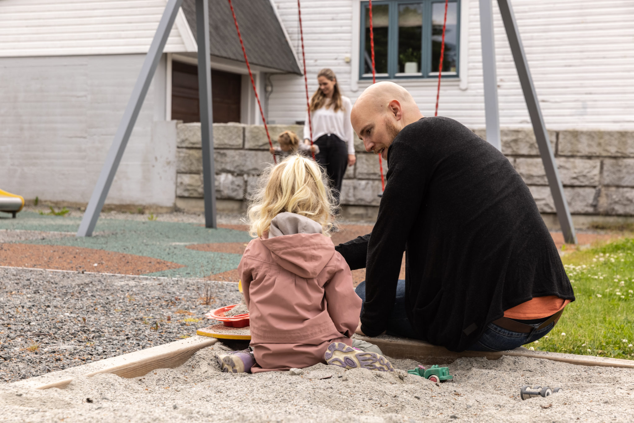 barn og en voksen i sandkasse, mens en kvinne står ved en huske og hjelper barnet