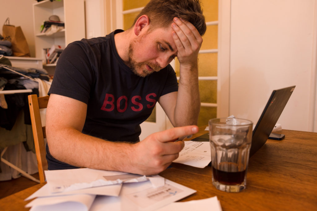 Mann sitter ved laptopen sin. Det er mange konvolutter og fakturaer. Han tar hånden til pannen og er frustrert