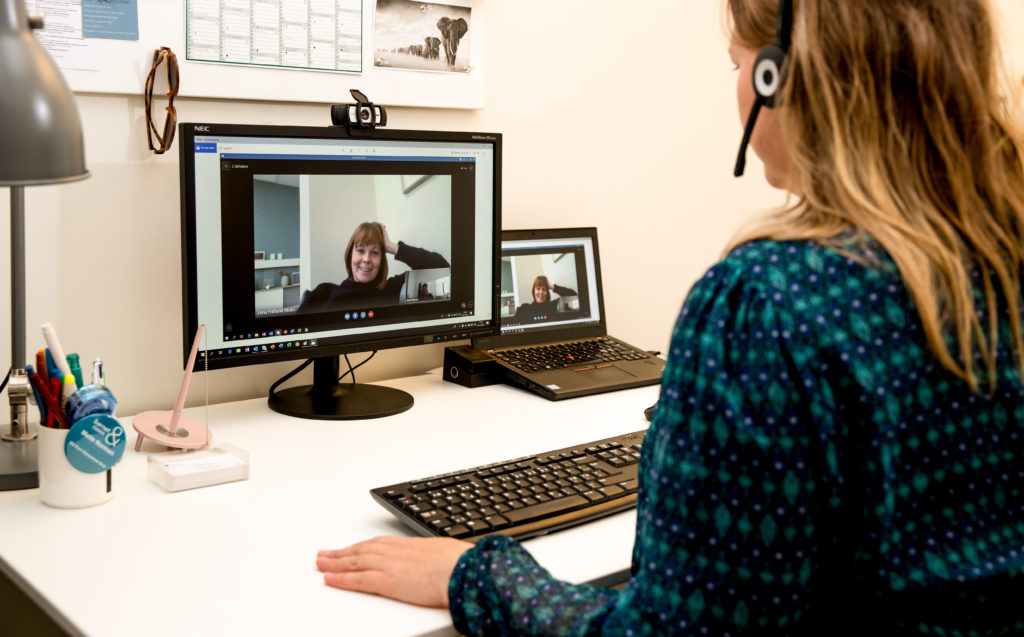 En terapeut sine foran PC og har en samtale med en bruker. Hun har headset på og smiler mot skjermen.