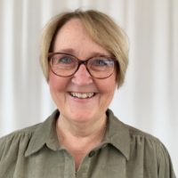 En kvinne i grønn bluse. Hun smiler og har på seg briller.