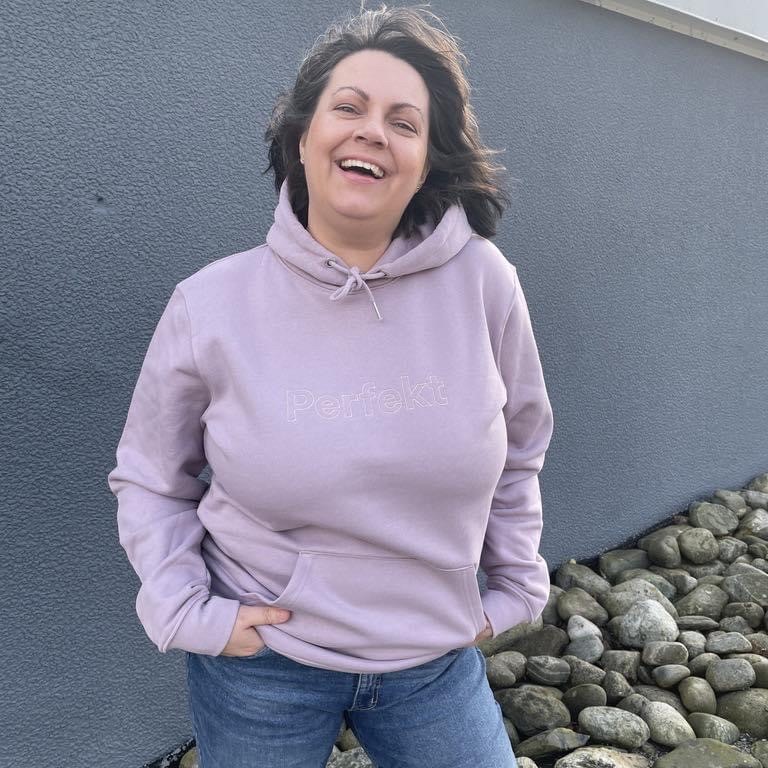 En kvinne står med lilla hettegenser med hendene i lomma og ler